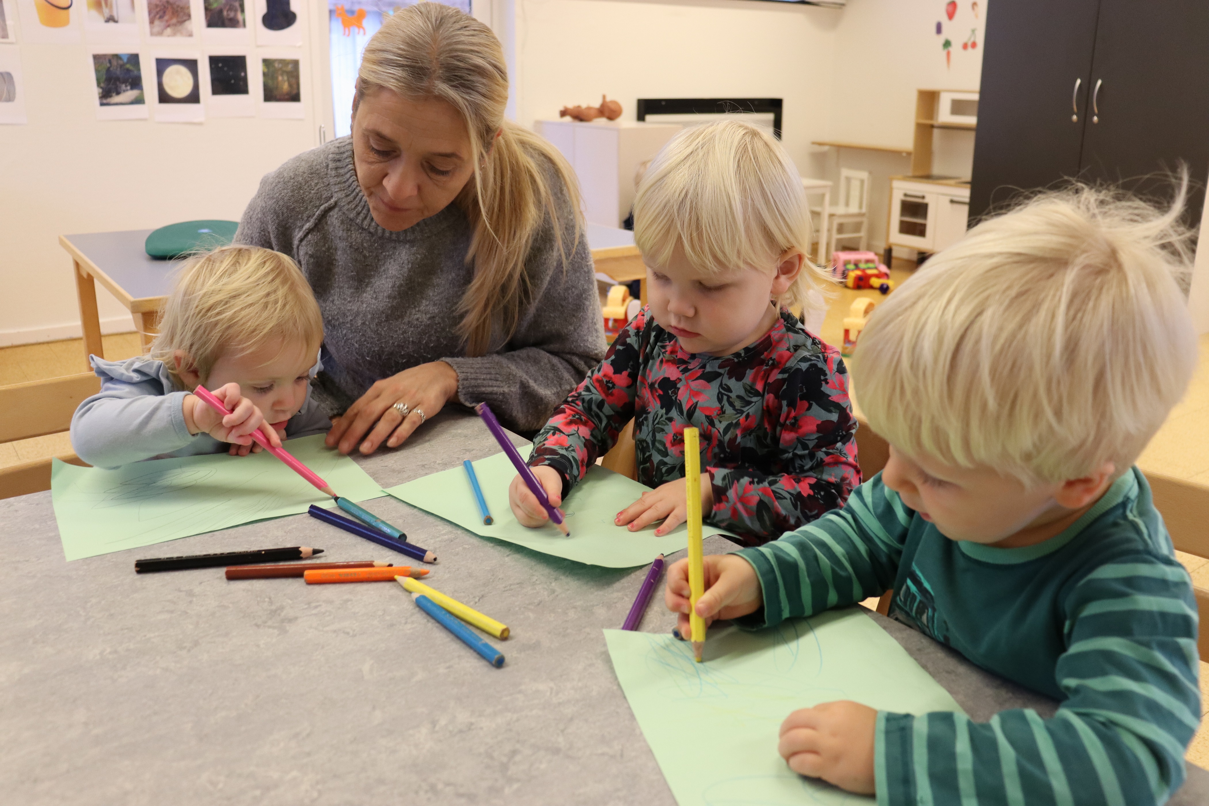 Pædagog sidder med barn og hjælper med at farve med farveblyanter, hvor der også sidder andre børn og farver